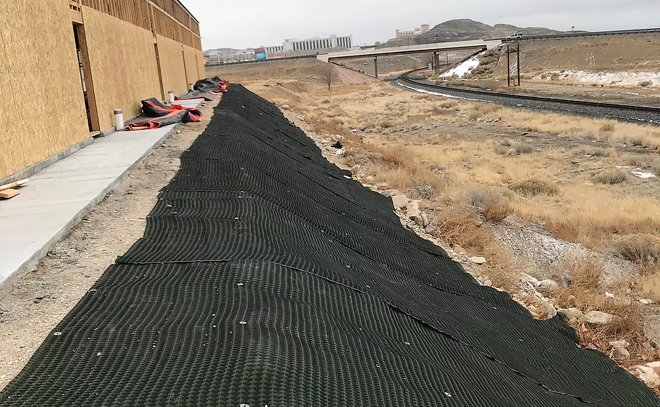 Sloped landscape with black erosion control matting, adjacent to a construction site. The scene is dry with sparse vegetation and overcast skies.