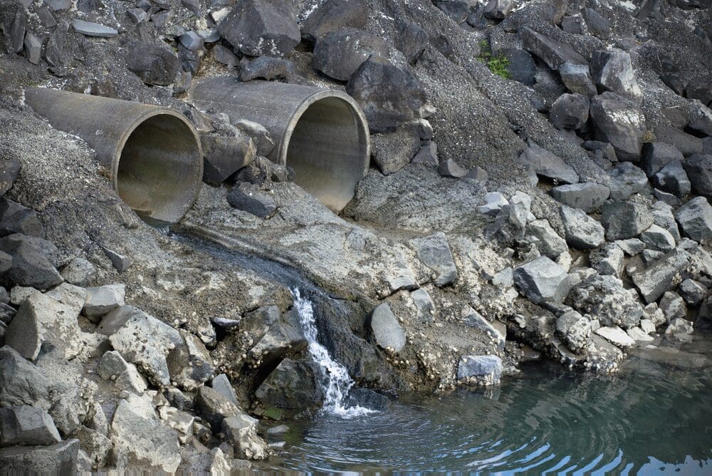 Two large concrete pipes release water onto rocky terrain, creating a small cascade into a calm body of water. The scene appears industrial.