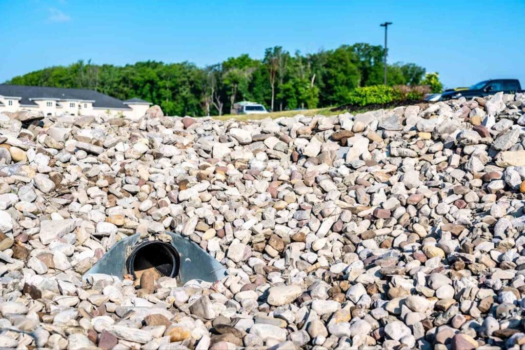 A pile of rocks with a drainage pipe opening, set against a backdrop of buildings and trees.