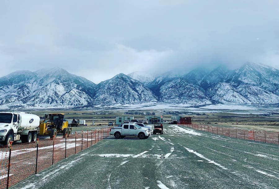 A snowy landscape with trucks and construction vehicles parked along a gravel road, set against a backdrop of rocky mountains and cloudy skies.