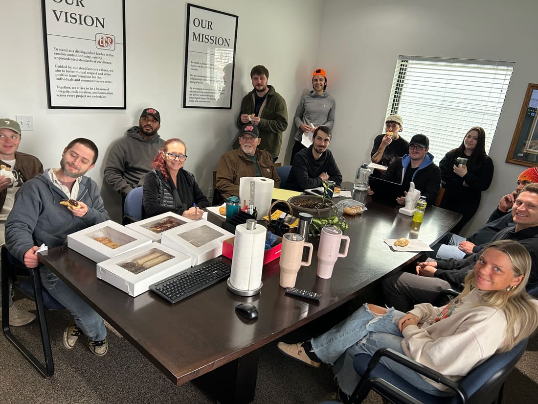 A group meeting in an office setting, with people seated around a table enjoying snacks and drinks, surrounded by motivational posters.