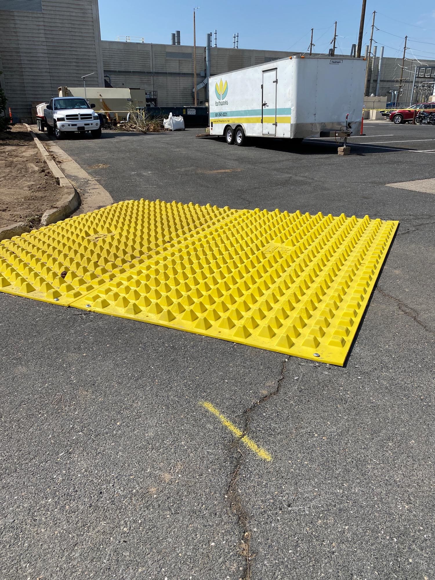 A bright yellow textured mat rests on asphalt, with a trailer and parked truck in the background under clear blue skies.