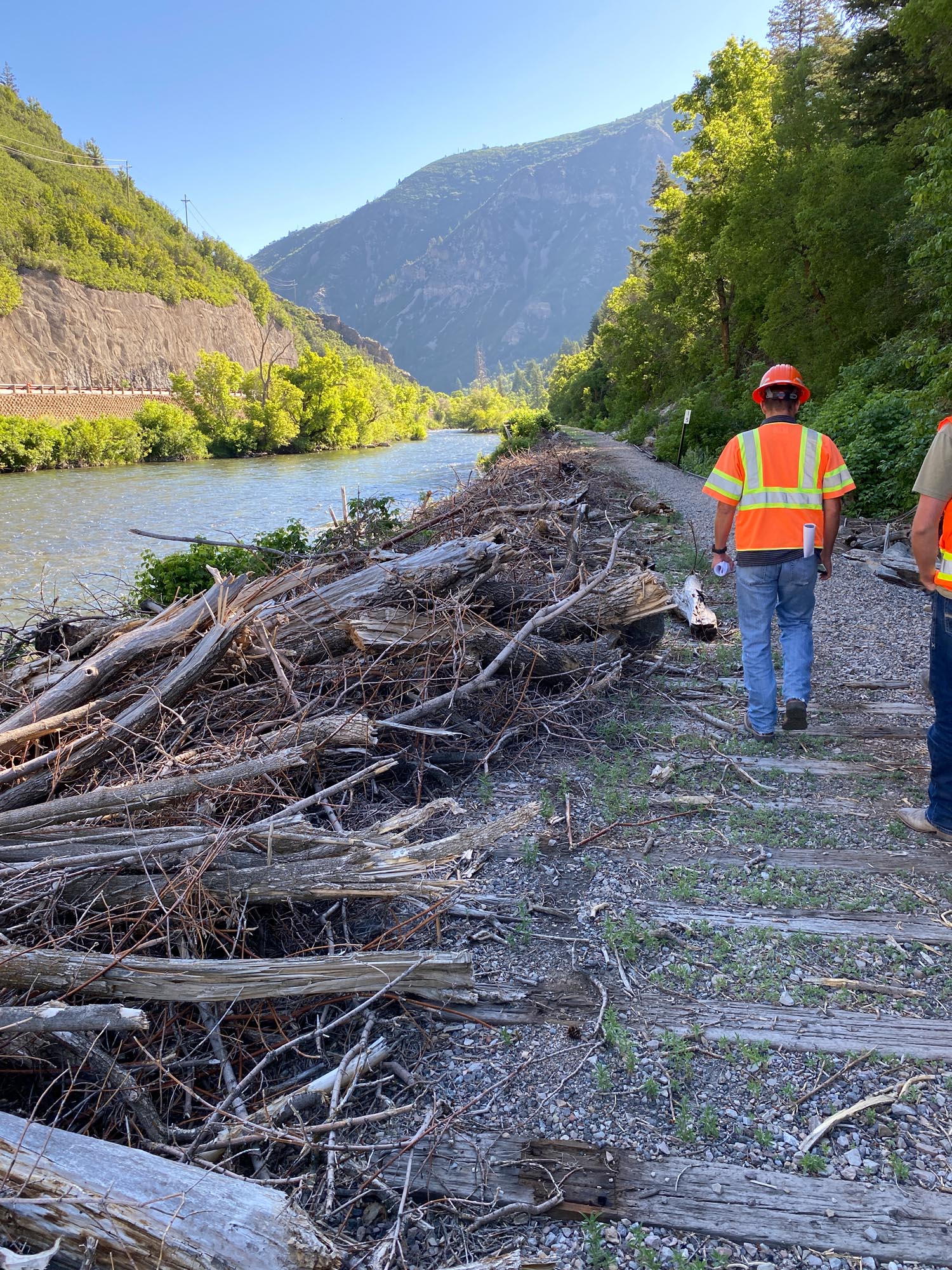 Two workers in safety gear walk along a riverbank lined with debris, surrounded by lush greenery and steep mountains under a clear sky.