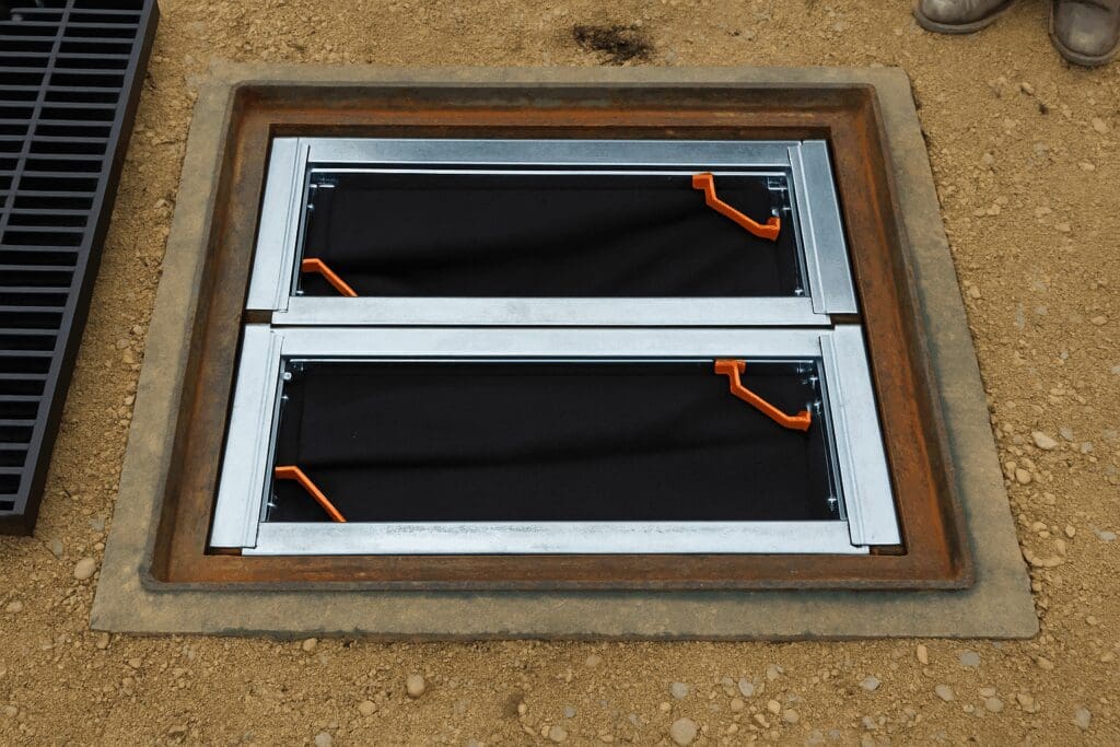 Photo of an open storm drain with two black filter screens and orange handles, surrounded by gravel and a grate.