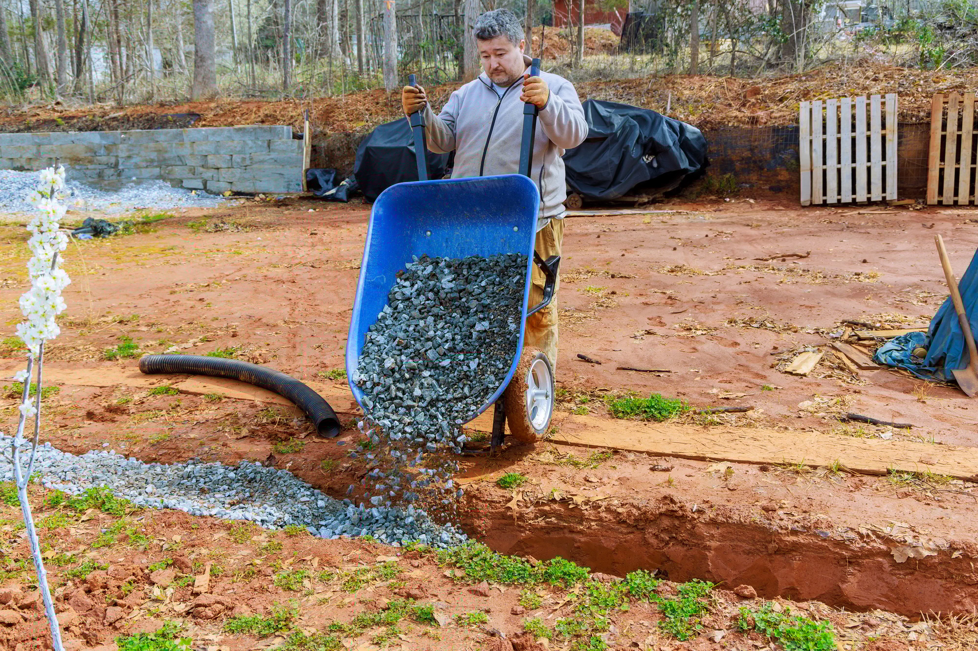 In wheelbarrow, a worker fills drainage pipes with crushed stone
