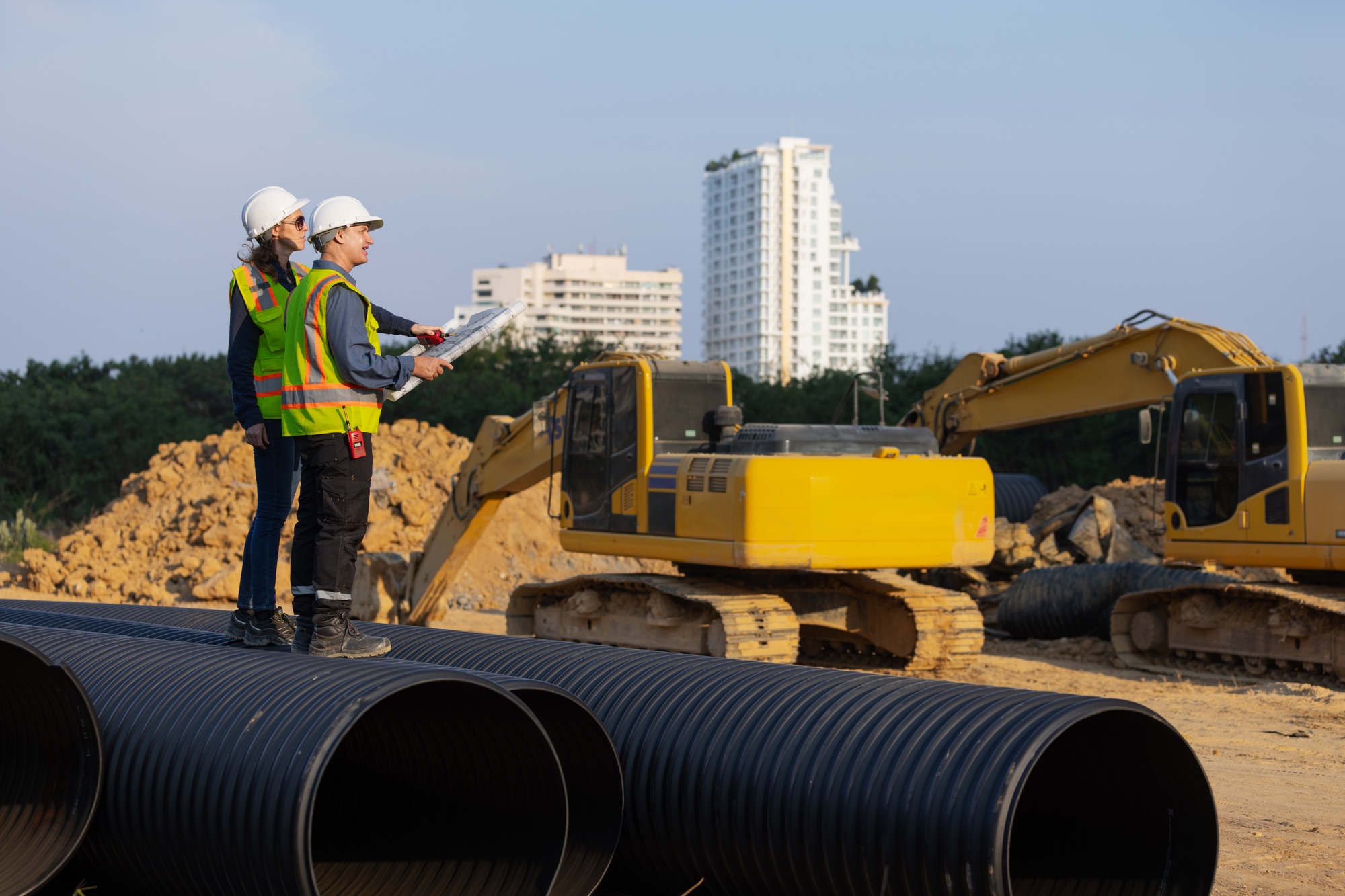 Two construction engineers in safety gear stand on large drainage pipes