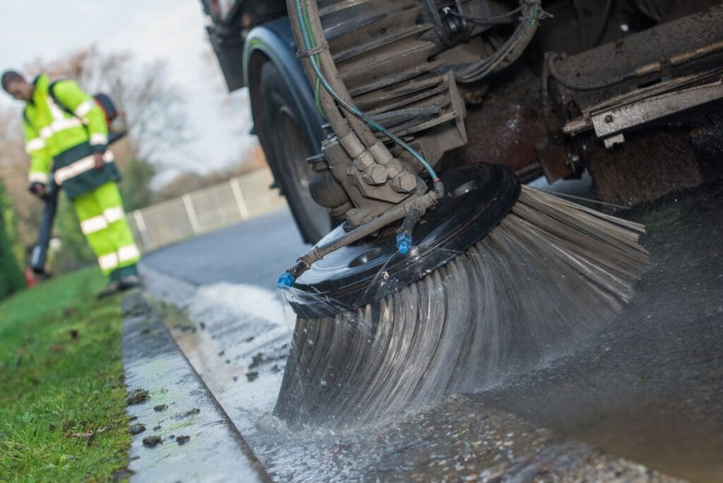 Street sweeper cleaning a wet road with a worker in a high-visibility vest nearby.